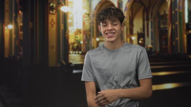 Teenage man in gray tee smiles with arms folded, right hand gripping left elbow, standing in ornate church building nave with pews and arches; confidence youthful ease.