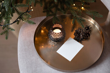Blank paper card mockup, candle and pine cone on cooper tray, Christmas tree shining lights, aesthetic holiday background for business branding, top view, copy space