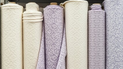Rolls of patterned fabric for upholstery and interior design. Assortment of cream and purple textiles on shelf in store. Home decor and sewing material selection