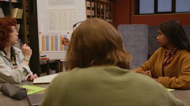 Black male English teacher with glasses explaining new grammar rule guiding diverse group of students through verb tense forms in language training course while sitting at table in library