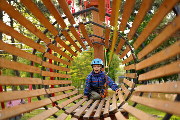 Excited young boy wearing a helmet and harness crawls through a wooden barrel obstacle on a high ropes adventure course.