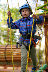 Young boy in a helmet and harness carefully walks across a narrow, suspended rope bridge on an outdoor adventure course.