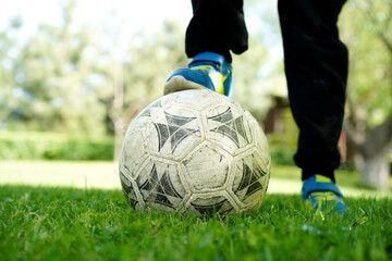 Close-up of a worn soccer ball on grass with a person’s foot resting on it outdoors.