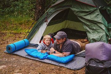 Father and smiling young son relaxing together at the entrance of a green tent in a shady forest campsite with camping gear.