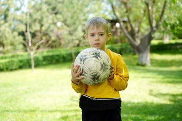  Young boy holding an old soccer ball while standing on green grass in a sunny park.