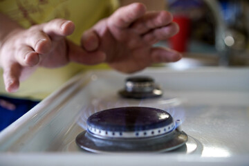 Close-up of a blue gas flame on a kitchen stove with blurred hands warming them above the burner, focused on home cooking.