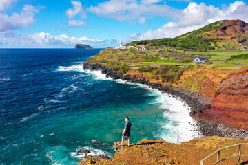 Aerial view of azure waters crash against the rugged coastline near a solitary figure gazing towards a distant island and the Ponta da Barca Lighthouse, Judeu, Azores, Portugal.