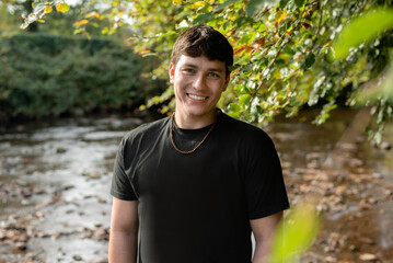 A smiling man in a black T-shirt stands by a leafy riverbank. A guy is happy after active recreation, running in park on a treadmill