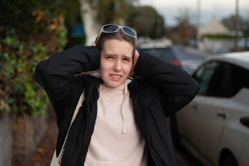 A young girl stands on a city street in a beige hoodie, black jacket. She holds her head with both hands, showing stress, frustration in urban setting. A girl is afraid of the noise of cars