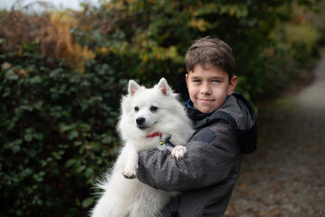A young boy hugs a white fluffy dog along autumn garden path. A boy is always happy when he walks with his dog, friendship between people and pets