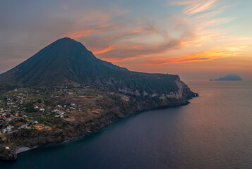 Aerial view of the dramatic coastline where dark cliffs meet the serene blue sea under a vibrant sky painted with orange and pink hues, Salina, Sicilia, Italy.