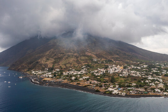 Aerial view of the dramatic dark volcanic slopes meeting the clustered buildings of the town against the backdrop of swirling clouds, Stromboli, Sicily, Italy.