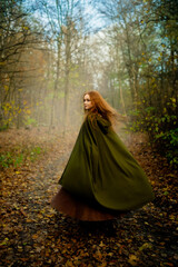 Naklejka premium Red-haired woman in a medieval green cloak and corset posing in a mystical mossy forest, evoking an elven fairy-tale atmosphere with ethereal light and magical fantasy vibes. 