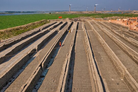 Aerial view of freshly laid bricks drying under the warm sun, contrasting with the distant green fields and wind turbines, Aminbazar, Dhaka Division, Bangladesh.