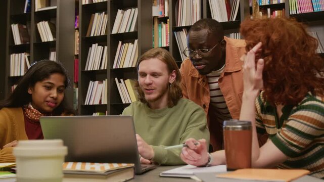 Multi ethnic group of smiling college students discussing homework assignment browsing information on laptop while studying together in library