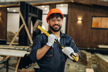 Big hammer is in hands. Portrait of a male carpenter posing in a woodworking workshop