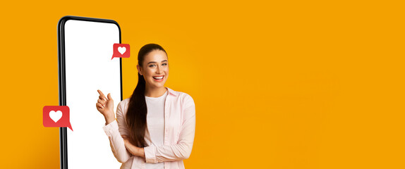 A cheerful woman stands beside a large phone screen, smiling and pointing at it. The background is...