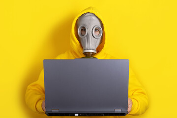 Individual in yellow hoodie and gas mask using laptop on yellow background. Concept of online security, protection, or isolation.