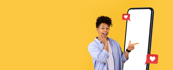 A young man stands next to a large smartphone screen, smiling and pointing with enthusiasm. The vibrant yellow backdrop enhances the lively mood, creating a fun atmosphere for digital engagement.