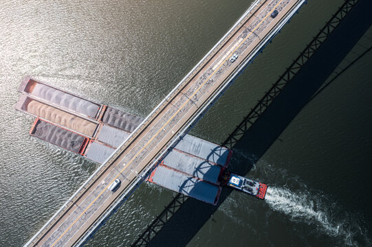 Aerial view of a barge navigating the Hudson River under the Bear Mountain Bridge, a stark contrast of industrial transport against the natural waterway, Bear Mountain, New York, United States.
