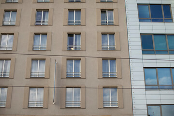 Beige hotel windows grid, single lit. Repetitive square frames with metal railings, white curtains, adjacent reflective glass