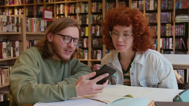 Cheerful young man and woman watching hilarious video on smartphone at library table while having break from study session