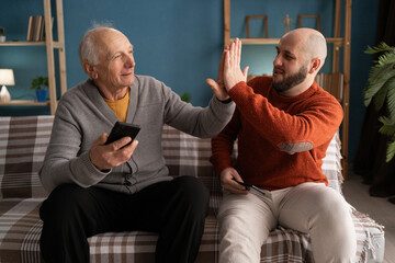 Senior elderly Father and adult son play video games with smartphone and have fun at home spending time together