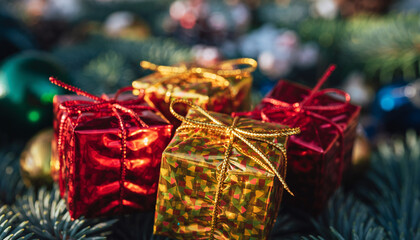 Close-up of small, shiny red and gold gift boxes with bows, nestled in the branches of a pine tree. Festive Christmas holiday background with bokeh lights and colorful ornaments.