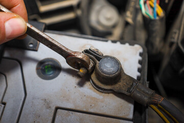 ​A close-up shot shows the process of unscrewing or tightening a nut on a car battery terminal using a wrench held by a hand. Signs of use, oxidation, and wires in the engine bay are visible.