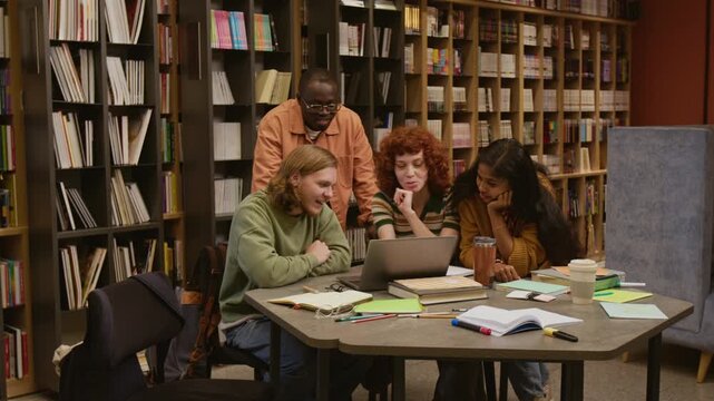 Group of laughing multiethnic college students watching video on laptop at library table while studying together after classes