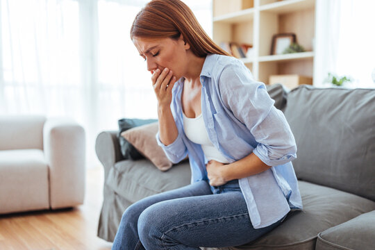 Woman Sits on Couch at Home Suffering Nausea and Stomach Discomfort, Gripping Her Abdomen