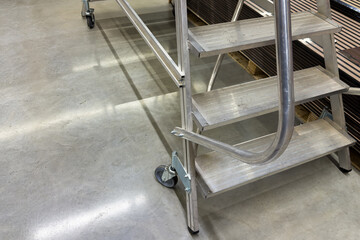 A set of rolling steps is positioned on a concrete floor in a workshop. The steps have a sturdy handrail and wheels for easy mobility. Workers use them to reach high areas safely during projects.