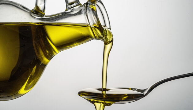 Olive oil pouring from a glass bottle into a metal tablespoon on a white background.