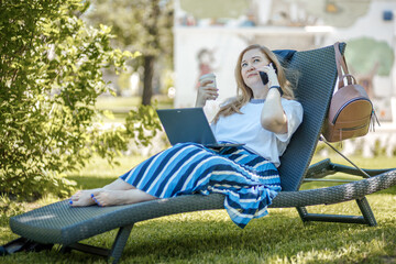 A middle-aged blonde woman with a laptop works in the garden on a sun lounger, a green home office concept.