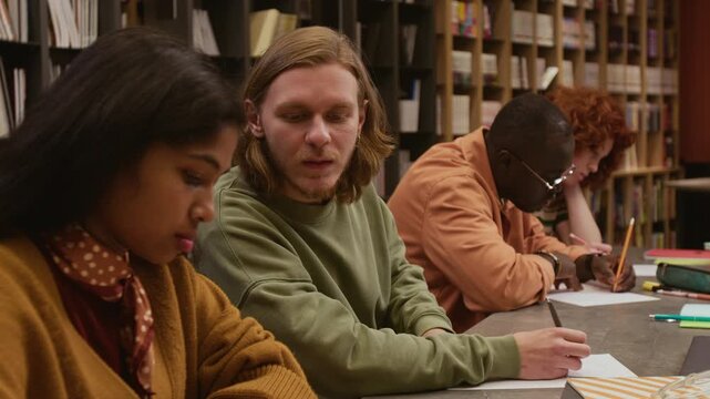 Side view of adult man cheating on examination test looking up answers on groupmates worksheet while sitting in row with multi ethnic group of students in language training course in library