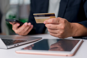 Close-up of hands holding a gold credit card and using a smartphone for online shopping. E-commerce concept with laptop and tablet on the desk.