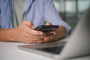 Close-up of a young man's hands holding a smartphone and typing a message on social media. Casual male using a mobile phone app at a desk with a laptop.