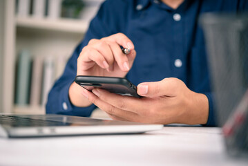 Close up businessman or freelancer hand holding a smartphone and a pen while sitting at a white desk with a laptop. checking email, mobile banking app, or social media for business marketing.