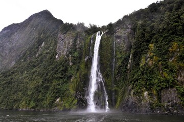 Majestic waterfall cascading down a steep green cliff into the sea in Milford Sound, New Zealand, showcasing the power of nature.