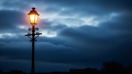 Vintage street lamp glowing with warm yellow light against a dramatic dark blue cloudy sky. Antique lantern post illuminating the city during a moody evening or stormy night.