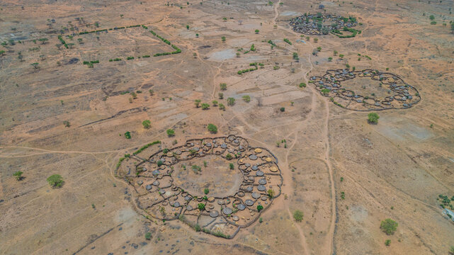 Aerial view of circular Manyatta villages surrounded by arid, ochre landscape, creating a stark contrast between human settlement and the vast African terrain, Karamoja, Northern Region, Uganda.