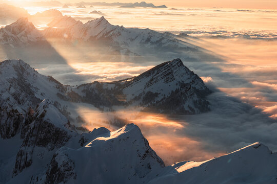 Aerial view of sunlight piercing through the clouds over snow-capped mountains, creating a dramatic contrast of light and shadow on the peaks, Wildhaus-Alt Saint Johann, St. Gallen, Switzerland.