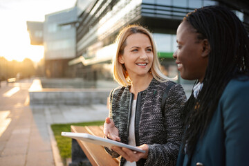 Two businesswomen discussing outdoors, using a digital tablet for work