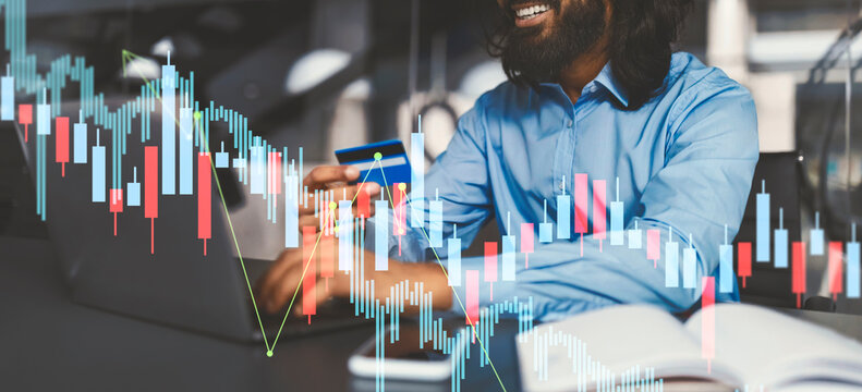 A professional man in a blue shirt smiles while shopping online with a credit card at his desk. Financial graphs are visible, creating a busy office environment that enhances the scene. - Powered by Adobe