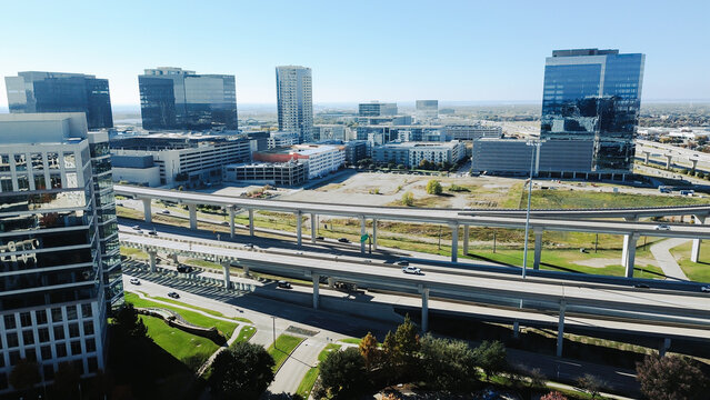 Multi-level interchange looping ramps and directional traffic, office towers, parking decks, landscaped buffers. Captures dynamic vehicular movement, urban engineering precision, Plano, Texas