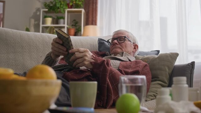 Handheld shot of drowsy elderly Caucasian man with cold taking nap while resting on sofa with book at home