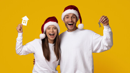 A joyful couple wearing Santa hats smiles widely while holding a toy house and keys. Their...