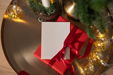 Christmas holiday postcard mockup with empty copy space. Blank paper card, red gift box on golden tray background with fir tree branch and shiny garland lights, festive present card design template