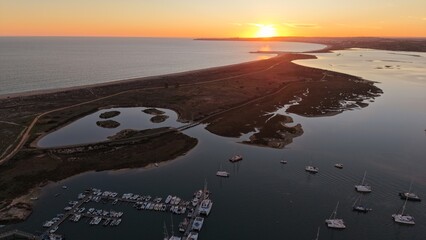 A sunset aerial view of a coastal lagoon with moored boats, winding waterways, reflective ponds, and a glowing horizon over the open sea.