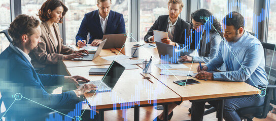 A group of five diverse professionals collaborates at a large table in a bright office. They focus on their laptops while enjoying a view of the city skyline through large windows.